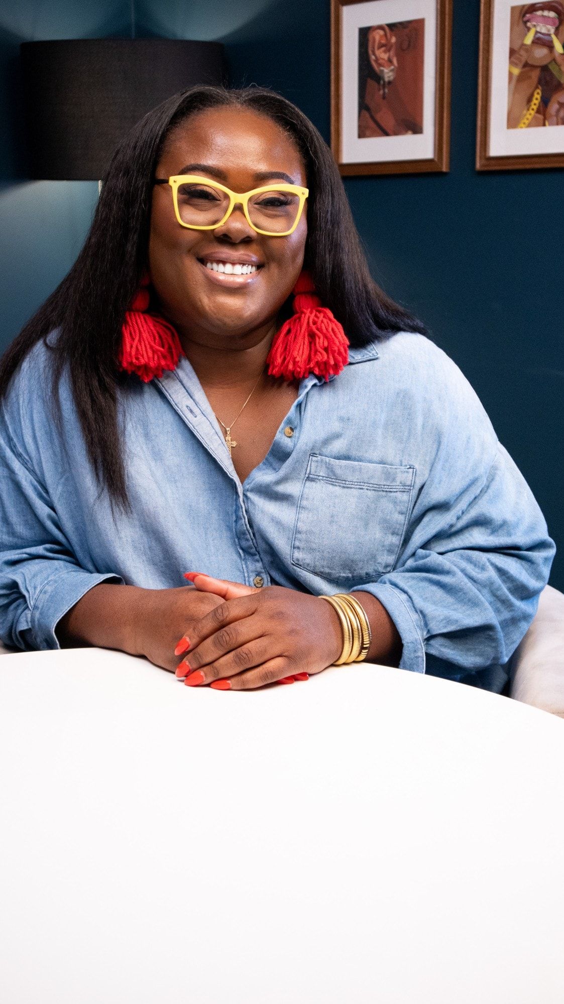 Ona Oghogho smiling at a table in a denim shirt, yellow glasses and red tassel earrings